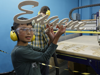 A child wearing safety glasses and headphones holds up a cut-out piece of wood spelling the word "Eugene".