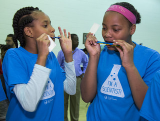 Two girls wearing "I'm a Scientist" t-shirts each blowing into a handheld sound machine experiment