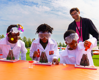 Three children wearing lab coats and goggles make volcanoes while a team member watches behind them.